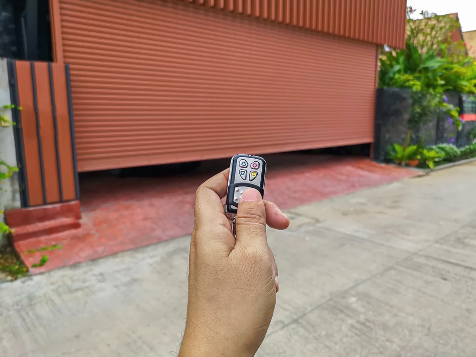 Scène extérieure résidentielle avec personne tenant une télécommande de portail automatique face à un garage fermé avec porte sectionnelle marron
