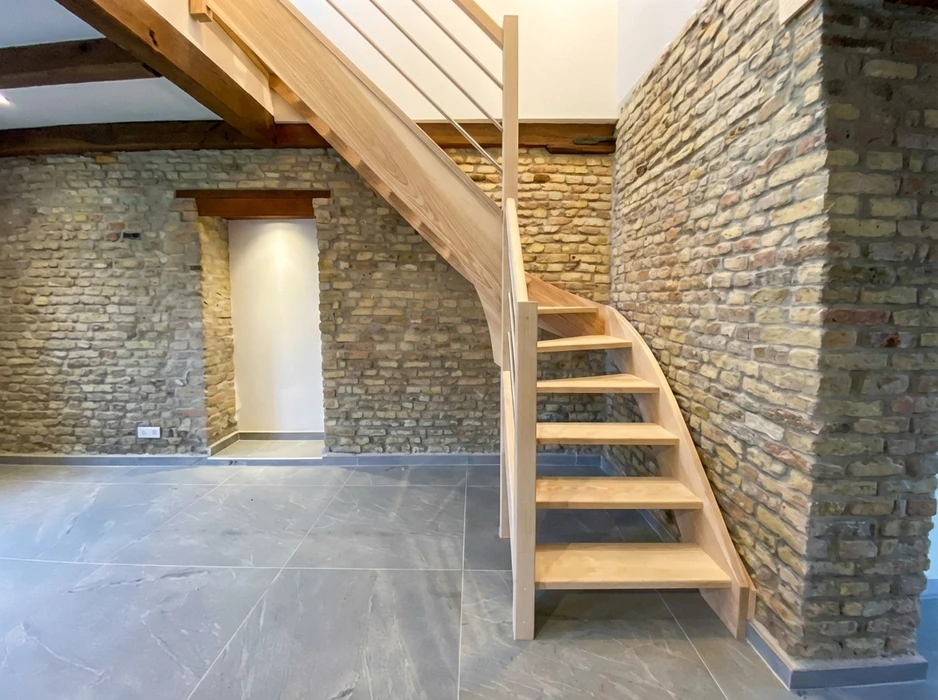 Façade en briques rouges d'un bâtiment résidentiel avec trois ouvertures : une fenêtre équipée d'un volet roulant métallique anthracite avec jardinière fleurie, une porte d'entrée en bois massif vertical naturel à arc surbaissé, et une petite porte métallique peinte noire